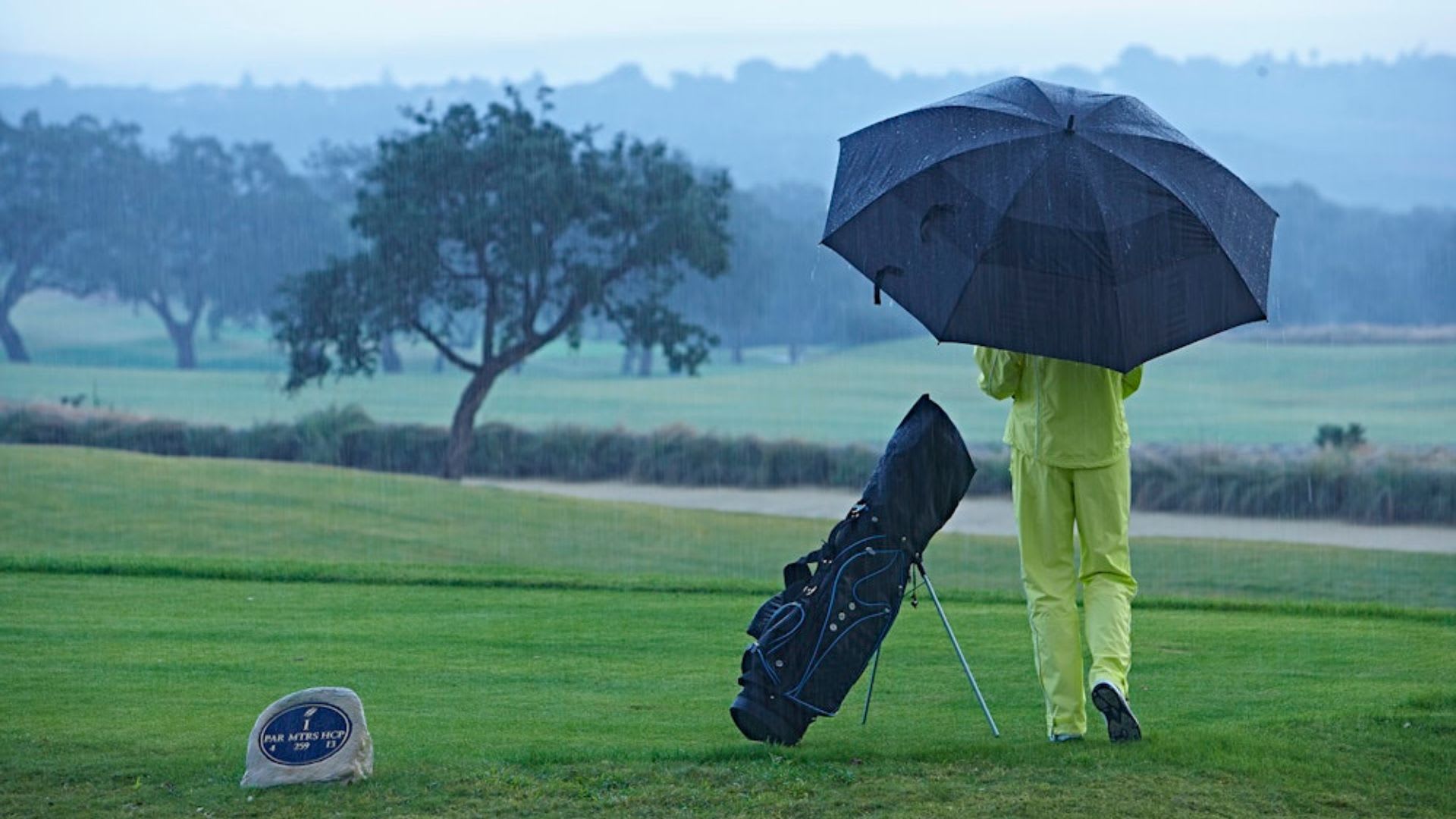 Person in yellow rain gear holding an umbrella and standing next to a golf bag on a rainy day on a golf course.
